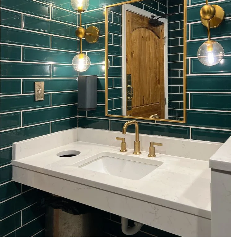 Bathroom vanity with green wall tile, white countertop, and brass fixtures